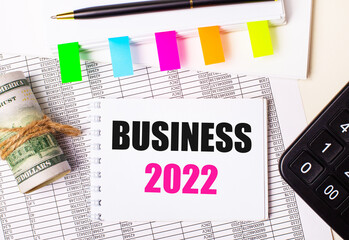 On a light background, a pen, dollars, a notebook with the text BUSINESS 2022 and bright stickers. Business concept