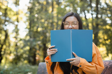 cheerful asian student in eyeglasses obscuring face with copy book in park