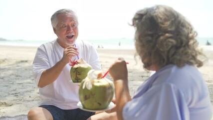 Asian family senior couple sitting on blanket have picnic drinking coconut juice together on the beach. Healthy elderly husband and wife relax and enjoy outdoor lifestyle activity on summer vacation - Powered by Adobe