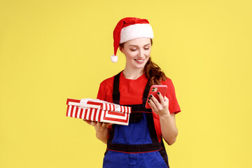 Positive delivery woman holding present box and using smart phone, specifies delivery address, wearing blue overalls and santa claus hat. Indoor studio shot isolated on yellow background. - obrazy, fototapety, plakaty
