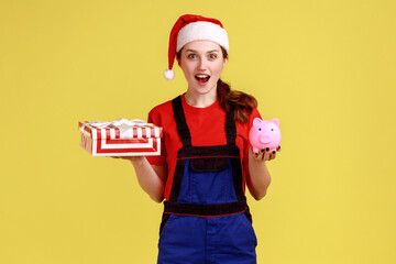 Courier girl holds wrapped Christmas gift and piggy bank, save money ordering delivery, advertising, wearing blue overalls and santa claus hat. Indoor studio shot isolated on yellow background.