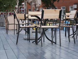 An open-air cafe is preparing to receive guests. New furniture for the cafe is installed. Tables and chairs made of black metal and light wood on a dark gray floor.