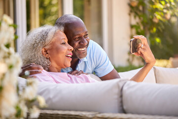 Senior Retired Couple Sitting Outside In Garden At Home Making Video Call On Mobile Phone