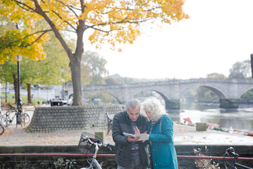 Senior couple with bicycles traveling, looking at guidebook along autumn river