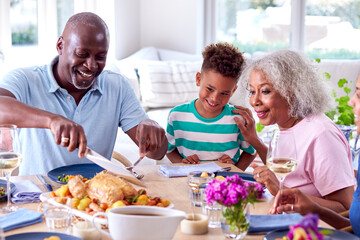 Grandfather Carving As Multi Generation Family Sit Around Table At Home And Enjoy Eating Meal