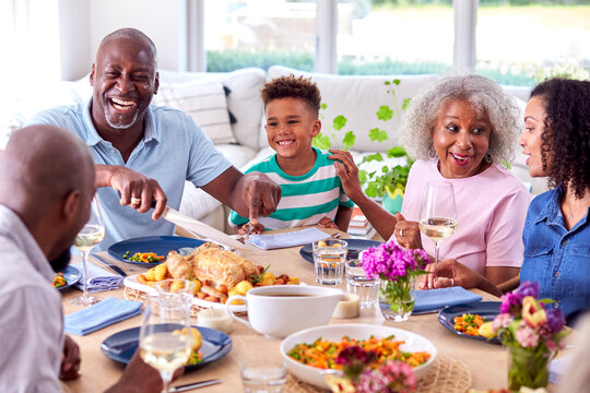 Grandfather Carving As Multi Generation Family Sit Around Table At Home And Enjoy Eating Meal