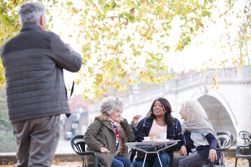 Senior man photographing active senior women friends at autumn park cafe