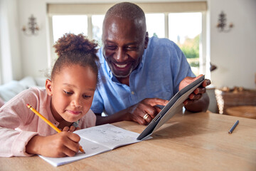 Grandfather Helping Granddaughter With Home Schooling Sitting At Table With Digital Tablet