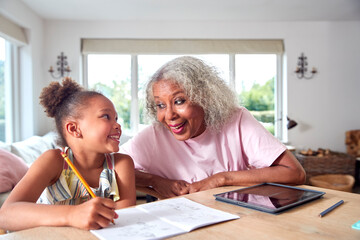 Grandmother Helping Granddaughter With Home Schooling Sitting At Table With Digital Tablet