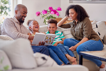 Parents Sitting On Sofa With Children At Home Reading Book Together