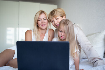 little girl with mom and grandmother are sitting on the bed in front of a laptop