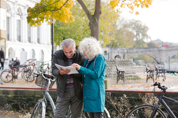 Senior couple with bicycles traveling, looking at guidebook along autumn river