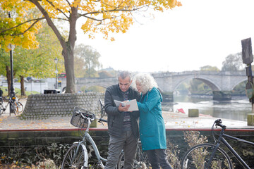 Senior couple with bicycles traveling, looking at guidebook along autumn river