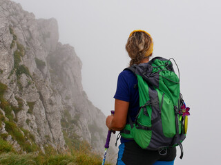 Photo of a Caucasian blonde hiker woman with green backpack, dressed in blue with yellow scarf hiking in a mountainous area with fog