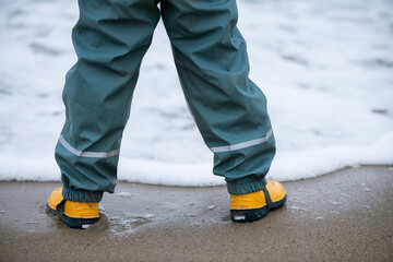 Kleinkind mit gelben Gummi Stiefel und Regenschutz Hose
steht am Meeres Strand vor schäumendem...