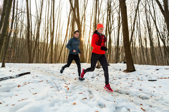 Man and Woman Running Together Through Woods in Winter. Smiling Couple Running Along Trail Through Forest on Cold Winter Day.