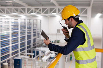 Male supervisor with digital tablet talking, using walkie-talkie on platform in factory