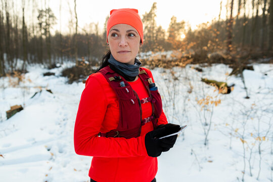 Female Runner Holding Mobile Looking for Way Out of Woods in Winter. Sportswoman Looking Around Forest Checking Location on Cellphone On Cold Winter Day.