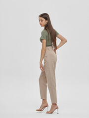Young elegant woman with long straight hair and natural make-up in green top and beige jeans standing and posing on bright grey background