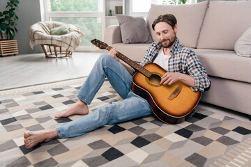 Photo of pretty handsome young man wear plaid shirt smiling sitting floor playing guitar indoors house room