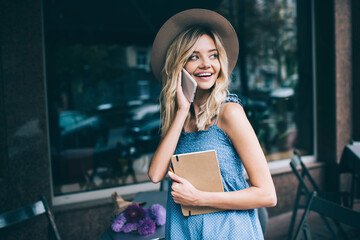 Cheerful hipster girl with education textbook calling to best friend for talking at street cafe, happy female student holding personal planner and smiling during positive international conversation