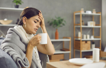 Sad unhappy exhausted sick woman suffering from common cold or flu fever, sitting on sofa wrapped in scarf, plaid and blanket, feeling unwell, touching forehead, holding mug and drinking warm tea