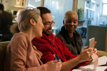 Three colleagues sitting on sofa, discussing paperwork, laughing