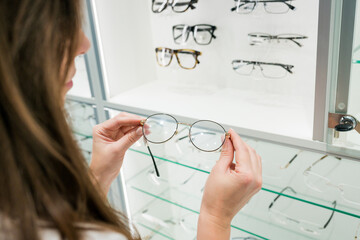 Close up of woman choosing glasses on eyewear display stand in optical store. Female taking frame...