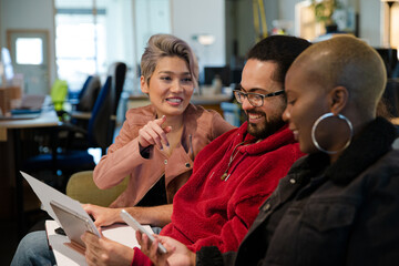 Three colleagues sitting on sofa, discussing paperwork, laughing