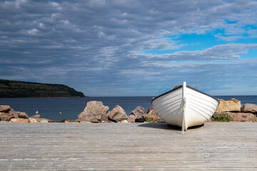 A white wooden fishing boat on the ocean shoreline under a blue sky with lots of clouds.  A land point is in the background meeting the horizon. The ocean is blue and calm with some gulls on rocks. - obrazy, fototapety, plakaty