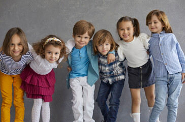 Excited positive children having fun standing in row smiling to camera and hugging against grey studio copy space. Cheerful classmates group portrait. Friendship and happy childhood concept