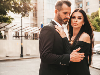 Beautiful fashion woman and her handsome elegant boyfriend in suit. Sexy brunette model in black evening dress. Fashionable couple posing in the street in Europe. Brutal man and his female outdoors