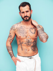 Fototapeta premium Portrait of handsome confident stylish hipster lambersexual model. Sexy modern man. Naked torso with tattoos.Fashion male posing in studio near light blue wall