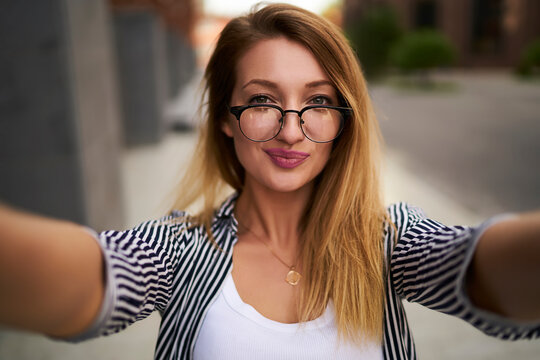 Close up portrait of attractive hipster girl in optical spectacles making selfie photos during leisure time at urban setting, beautiful woman in glasses photographing herself during free day outdoors