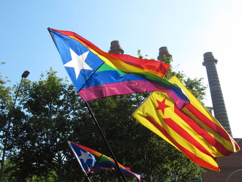 Two waving flags in a Pride Parade: independence catalonian flag and pride flag with white star in a blue triangle. Three background chimneys