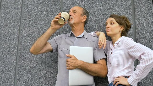 Young woman and senior man are waiting for a meeting standing near the grey wall outdoors in the morning. Colaboration between older coworker and younger colleage