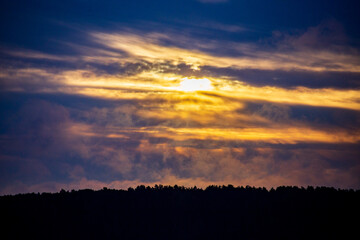 Fototapeta premium Evening river landscape with the setting sun. Calm water and dramatic sky with clouds. Trees grow on the nearest shore. There is a copy space
