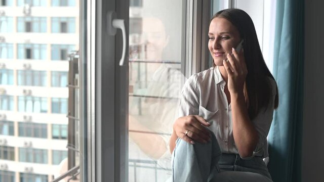 Charming woman talking on the smartphone sitting on the window cill at home. Smiling young woman chatting with friend on mobile phone, has pleasure phone conversation