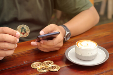 Obraz na płótnie Canvas Male hand holding a bitcoin while using phone with a cup of coffee on wooden table inside the cafe. Cryptocurrency trading concept
