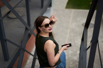 Portrait of pretty Caucaisan hipster girl in stylish sunglasses smiling at camera during leisure daytime in city, happy hipster girl with modern smartphone gadget for networking posing at urban stairs