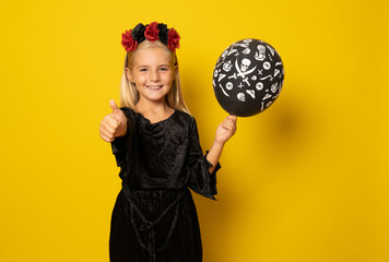 Fototapeta premium Portrait of little girl in black clothing holding black balloon with thumb up isolated over yellow background.