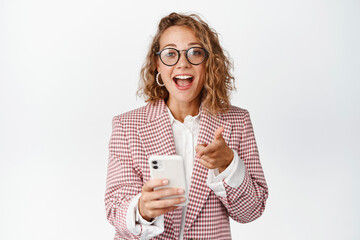 Surprised business woman holding mobile phone and pointing at screen, reaction to something awesome on smartphone, white background