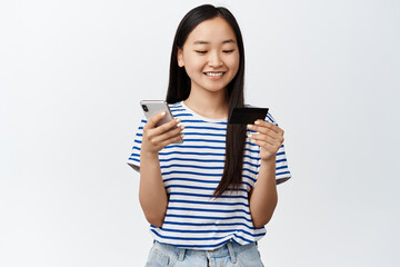 Online shopping. Smiling asian girl using mobile phone and credit card, paying in smartphone, standing over white background