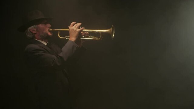 Bluesman plays trumpet in dark smoky room at spotlight, side view. Man in hat plays trumpet on black background