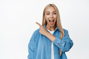 Excited blond woman smiling, pointing finger at upper left corner with amazed face, showing awesome...