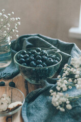 Fresh sweet blueberry in glass bowl with white flowers. Ripe berries for romantic breakfast under drizzling rain. Dark food concept	