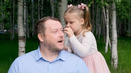 little daughter, 3 years old, tells her daddy in her ear and laughs. The concept of a happy childhood, fatherhood, parenting. A trusting relationship between parents and children. Fathers day