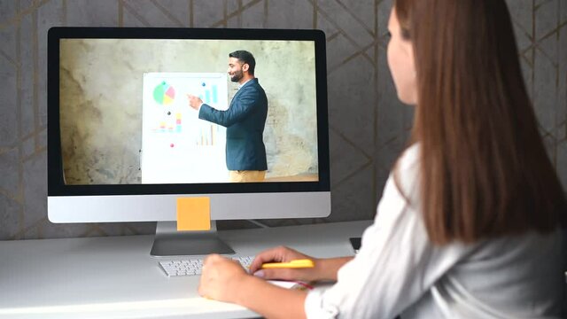 Indian male business coach is holding online training to young female student or employee. Intelligent businessman on the laptop screen involved virtual meeting, showing graph, woman takes notes