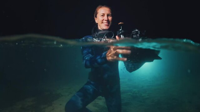 Underwater photographer in night lake. Young woman in wetsuit holds the camera in underwater housing and stands in the lake