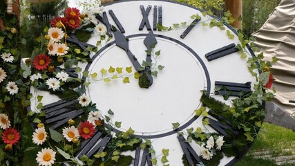 Large wall clock on the field in blooming flowers, showing the time 11:55, 12:00. time management concept, daylight saving time. Spring or wedding. Picnic for womens day